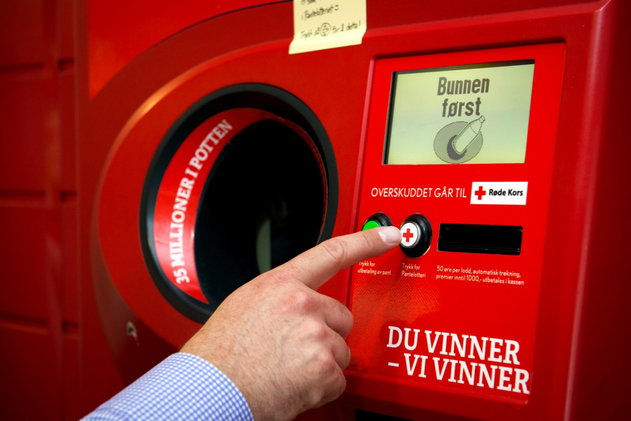 Foto eines Pfandlotterieautomaten in Norwegen. Abgebildet ist eine Hand, die auf einen Button drückt, um an der Lotterie zugunsten des Roten Kreuzes teilzunehmen.