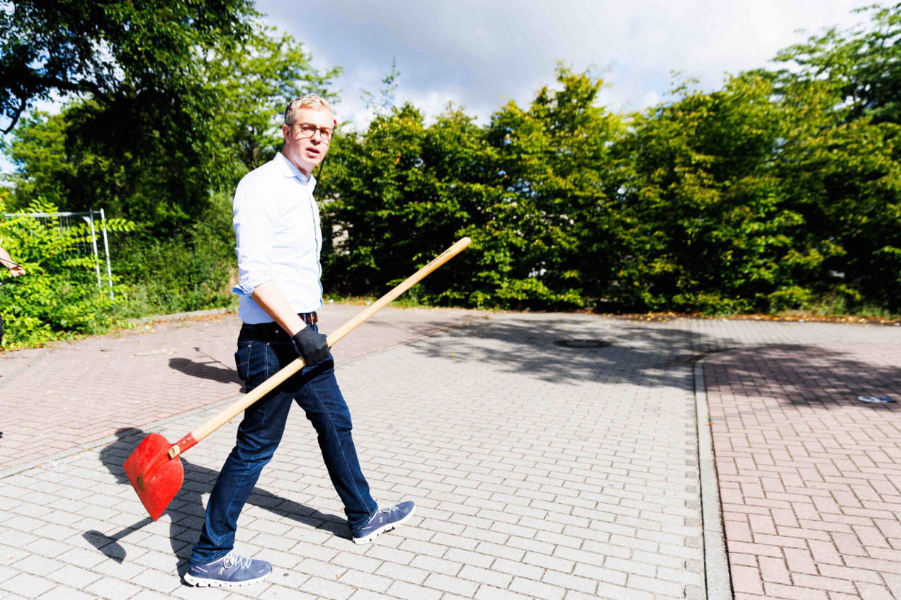Stefan Evers mit Hemd, Handschuhen und großer Schaufel, läuft über einen sonnigen Parkplatz